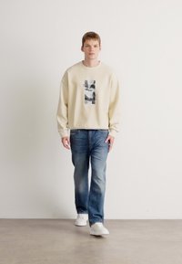 Beige hettegenser med grafisk trykk, kombinert med blå jeans og hvite joggesko. Passformen er avslappet med senkede skuldersømmer.