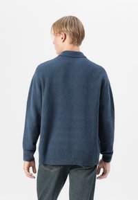 Suéter de punto azul marino oscuro con un acabado texturizado, de manga larga y con cuello. Llevado sobre unos jeans azules, mostrando un ajuste relajado.