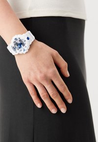 Montre en résine blanche avec des accents bleus et argentés, grand cadran rond, boutons texturés et bracelet en caoutchouc, portée sur une main partiellement visible.