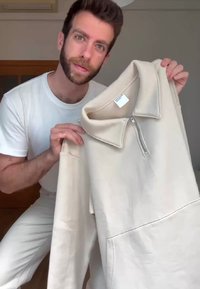 Beige sweatshirt med blixtlås i kragen, mjuk textur, långa ärmar och en framficka, hålls av en person i en ljus klädsel.