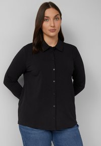 Zwarte blouse met lange mouwen en een kraag, voorzien van een knoopsluiting aan de voorkant en een relaxte pasvorm. Het materiaal lijkt zacht met een subtiele glans.