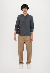 Dunkelgraues Hemd mit langen Ärmeln, beige weit geschnittene Hose und weiße Sneaker; eine kleine braune Umhängetasche hängt von der Schulter.