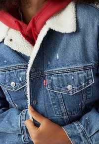 Džínová bunda s bílým ovčím límcem, opotřebovanými detaily a červenou n ášivkou Levi's. Obsahuje zapínací kapsy a klasickou modrou barvu.