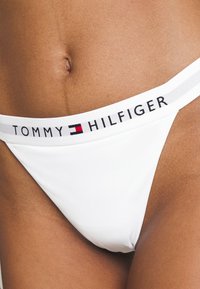 Slip in bikini bianco con una cintura elastica brandizzata con la scritta "TOMMY HILFIGER" in nero, rosso e blu. Tessuto liscio e copertura minima.