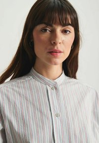 Chemise à bouton avec des rayures verticales en blanc, bleu et rouge. Dotée d'un col mandarin et d'un tissu lisse et texturé.