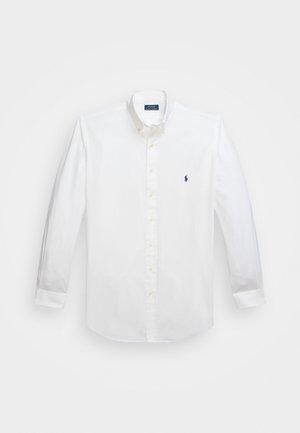Chemise blanche à manches longues en tissu coton, avec un col boutonnière, un logo bleu marine sur la poitrine et une fermeture par boutons. Texture lisse.