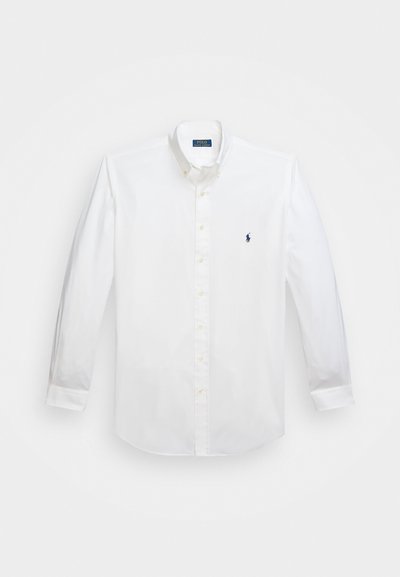 Chemise blanche à manches longues en tissu coton, avec un col boutonnière, un logo bleu marine sur la poitrine et une fermeture par boutons. Texture lisse.