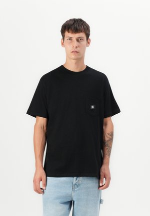 Črna bombažna majica s krožnim ovratnikom, kratkimi rokavi in žepom na levi strani prsi, ki vsebuje majhno logo. Model nosi svetlo modre jeans hlače.