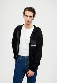 Sweatshirt preto com fecho, com bolso canguru e detalhe em logo branco, usado sobre uma t-shirt branca; combinado com jeans de denim azul.