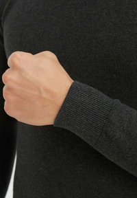 La manche d'un pull en tricot gris foncé présente un poignet à rayures. Une main est visible avec des veines définies, montrant la texture et la qualité du matériau.