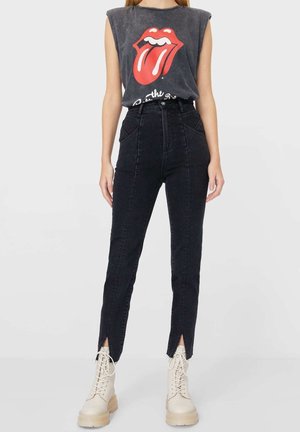 Person iført sort ærmeløs top med Rolling Stones-logo, sort højtaljede jeans med slidser ved anklerne foran og beige snørestøvler.