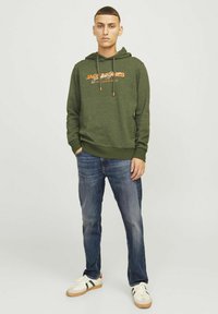 Sudadera verde con cordones y logotipo, combinada con jeans azules y zapatillas blancas con acentos en verde y rojo. Diseño simple y casual.