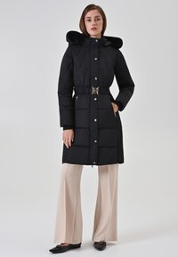 Zwarte doorgestikte pufferjas met een capuchon van nepbont, ingesnoerde taille, zilverkleurige knopen en zijzakken, gedragen met beige wijde pijpen broek en zwarte platte schoenen.