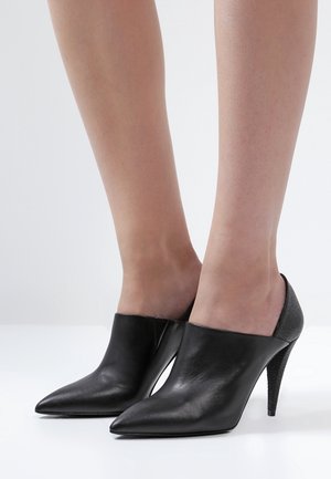 Mules de tacón alto de piel negra con punta afilada y diseño recortado. Un acento texturizado en el tacón añade detalle. Acabado suave en el cuerpo.