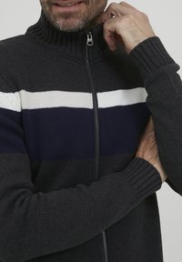 Pulover cu fermoar, tricotat, de culoare închisă, cu guler cu nervuri, având dungi orizontale în alb și bleumarin. Textură moale, mâneci ajustate.