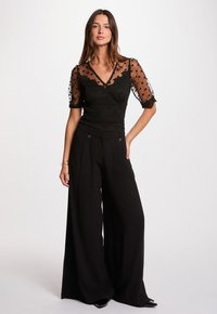 Zwarte kanten top met doorschijnende stippenmouwen, gecombineerd met zwarte wijde broekspijpen. De outfit heeft gedetailleerde afwerking en een v-hals.