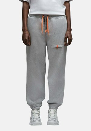 Personne portant un pantalon de survêtement gris avec des poignets élastiques, des cordons orange et noirs, une poche avant avec une étiquette orange, des baskets blanches et une chemise blanche.