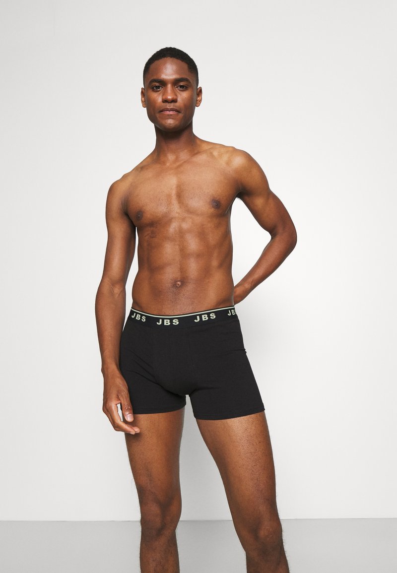Svarta boxerkalsonger med en mjuk textur och en elastisk midjebrand med "JBS" i kontrasterande grön text. Modellen poserar med en hand på höften.