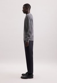 Maglione grigio lavorato a maglia con una texture a coste, abbinato a pantaloni scuri a gamba stretta e scarpe di pelle nera, visto di lato.