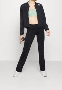 Veste bomber noire, brassière de sport verte, leggings noirs et baskets blanches. La veste est dotée d'une fermeture éclair et d'un logo Nike.