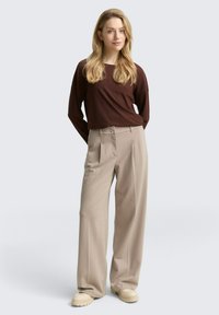 Vrouw met lang blond haar, gekleed in een donkerbruine lange mouw top, beige plooibroek en beige chunky schoenen, staand tegen een effen achtergrond.