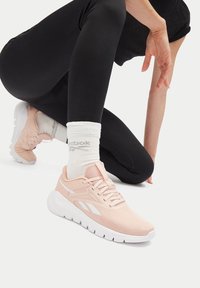 Osoba kľačí na jednom kolene v čiernych legínach, bielych ponožkách Reebok a svetloružových teniskách Reebok s bielymi podrážkami.