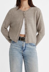 Cardigan marrone chiaro con scollatura rotonda, chiusura con bottoni frontali e orlo a coste. Indossato sopra una maglietta bianca e abbinato a jeans blu.