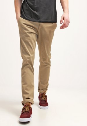 Mand går iført opkørte beige bukser, en sort skjorte og røde sneakers med brune snørebånd på en hvid baggrund.
