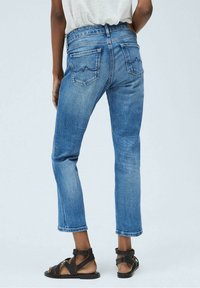 Vaqueros de mezclilla azul con un corte ajustado, dobladillo deshilachado y dos bolsillos traseros con un detalle de costura en zigzag. Llevados con sandalias negras de tiras.