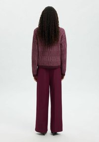 Une personne avec de longs cheveux bouclés se tient de dos, portant un pull mauve texturé et un pantalon bordeaux à jambes larges avec des talons noirs.