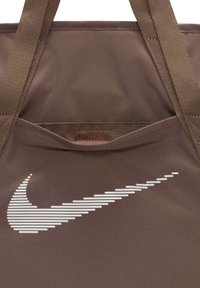 Bolsa tote marrón con una tela texturizada, que presenta un logo de Nike blanco y rayado en un lado y un pequeño bolsillo exterior. Incluye asas dobles.