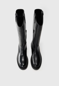 Une paire de bottes en cuir noires, longues, avec des bouts ronds et des fermetures éclair latérales, se tenant debout sur un fond blanc uni.