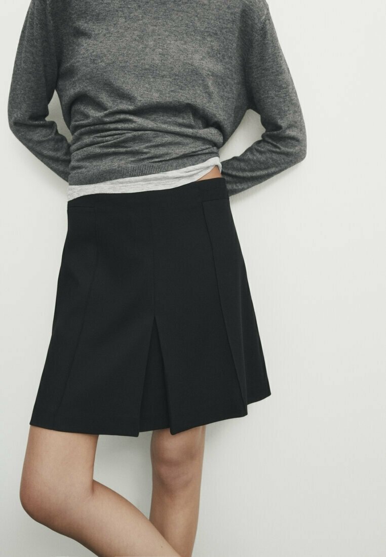 Personne portant une jupe plissée noire et un pull gris, debout devant un fond blanc uni avec les bras dans le dos.
