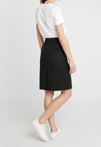 Svart knälång kjol med rak skärning, subtil textur, slits bak, och hög midja, kombinerad med en vit t-shirt och vita sneakers.