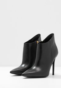 Bottes de cheville en cuir noir avec un bout pointu, un design épuré, une texture lisse et un fin talon haut. Elles présentent un détail de fermeture éclair dorée.