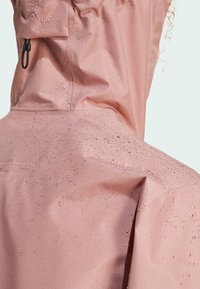 Giacca rosa impermeabile che mostra gocce su una superficie liscia e opaca. Tra le caratteristiche ci sono un cappuccio con coulisse e un design pulito e minimalista.