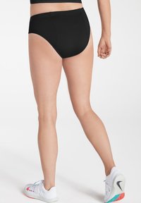 Une personne se tient de dos, portant des sous-vêtements noirs taille haute et des chaussures de sport blanches, sur un fond clair.