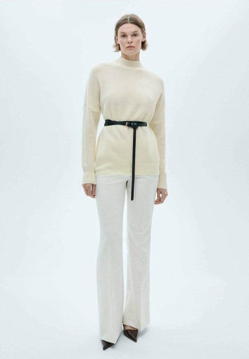 Maglione a maglia crema con collo alto, abbinato a pantaloni bianchi aderenti e una cintura nera. Le scarpe presentano un design a punta.