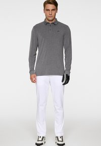 Mężczyzna w szarej koszulce polo z długim rękawem, białych spodniach, czarno-białych trampkach i czarnej rękawiczce golfowej na prawej ręce stoi na białym tle.