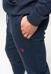 Marinblå joggare i mjukt tyg, med en sidoficka, elastisk midja och en liten röd broderad logotyp på låret.