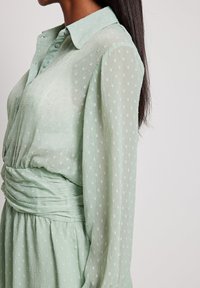 Abito verde chiaro con colletto a camicia, corpetto plissettato e maniche lunghe. Il tessuto presenta un leggero motivo testurizzato e un particolare vita arricciata.