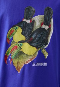 T-shirt bleu avec un graphique coloré de deux toucans perchés sur une feuille, avec le texte "ÉQUIPEMENT TOUTES CONDITIONS" en dessous.