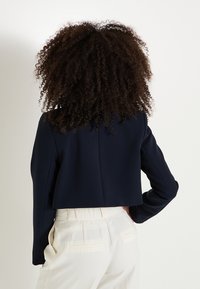 Veste cropped marine à manches longues, design structuré, texture lisse et coupe ajustée. Portée sur un pantalon de couleur claire.