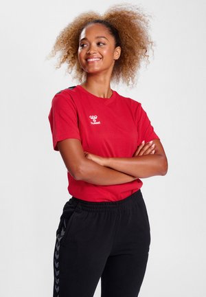 Rød t-shirt med korte ærmer og rund halsudskæring, prydet med en hvid logo på venstre bryst. Sammen med sorte bukser med elastik i taljen, der har sidelister.