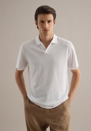 Giovane uomo visto frontalmente, indossa una polo bianca a maniche corte e pantaloni marrone chiaro, in piedi davanti a uno sfondo beige uniforme.