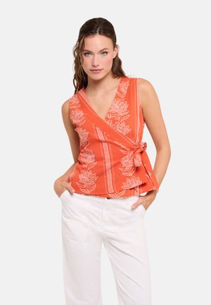 Mujer con top cruzado naranja sin mangas con patrones florales blancos y pantalones blancos, posando con las manos en los bolsillos delante de un fondo liso.