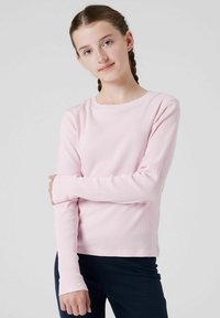 Giovane con capelli intrecciati che indossa una maglietta a maniche lunghe rosa chiaro e pantaloni scuri, in posa con un braccio incrociato sul torso.