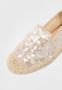 Bež espadrila s gornjim dijelom od šljokica, okruglim vrhom, teksturiranom jute potplatom i prošivenim detaljima oko ruba.