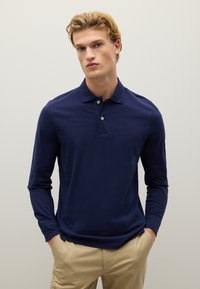 Langärmliges Navy-Polohemd mit weicher Textur, Drei-Knopf-Knopfleiste und dezenten Steppdetails, kombiniert mit hellen Hosen.