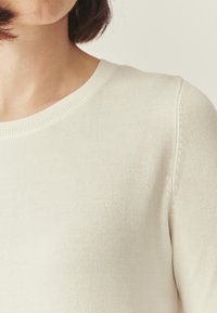 Pull léger crème avec un col rond, un col en maille côtelée et une texture tricotée lisse, avec des coutures discrètes aux épaules.